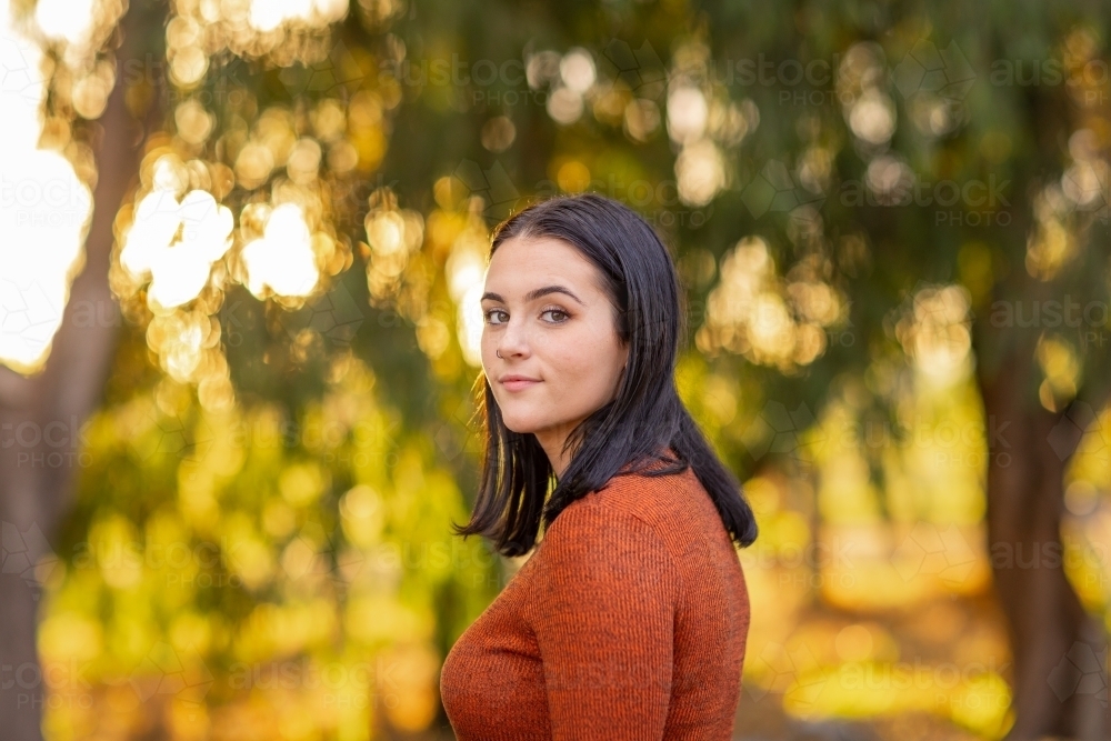 dark-haired teen girl looking over her shoulder against blurry background of trees - Australian Stock Image