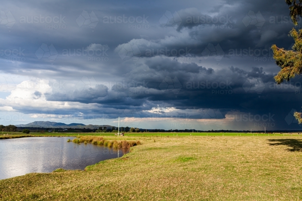 dark stormy rain clouds incoming over sunlit rural farm paddock and dam - Australian Stock Image