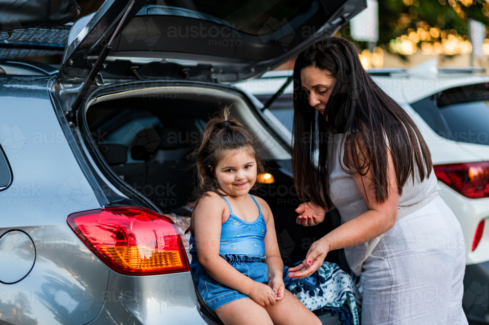 Daughter sits on the back of a car while mother talks to her in a parking area at dusk. - Australian Stock Image