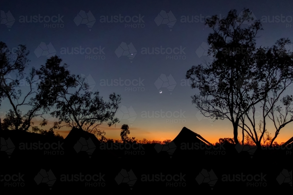Dawn breaking over safari tents with moon and stars in dark blue sky - Australian Stock Image