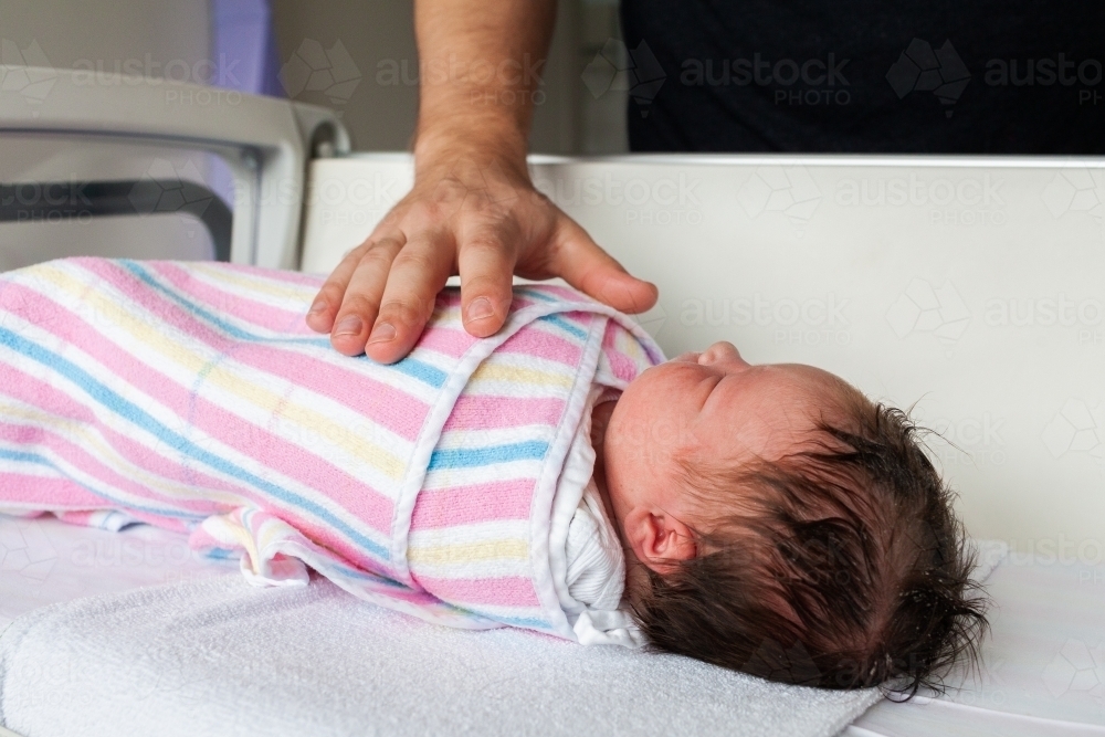 Day old newborn baby in hospital cot wrapped in blanket with dad's hand - Australian Stock Image