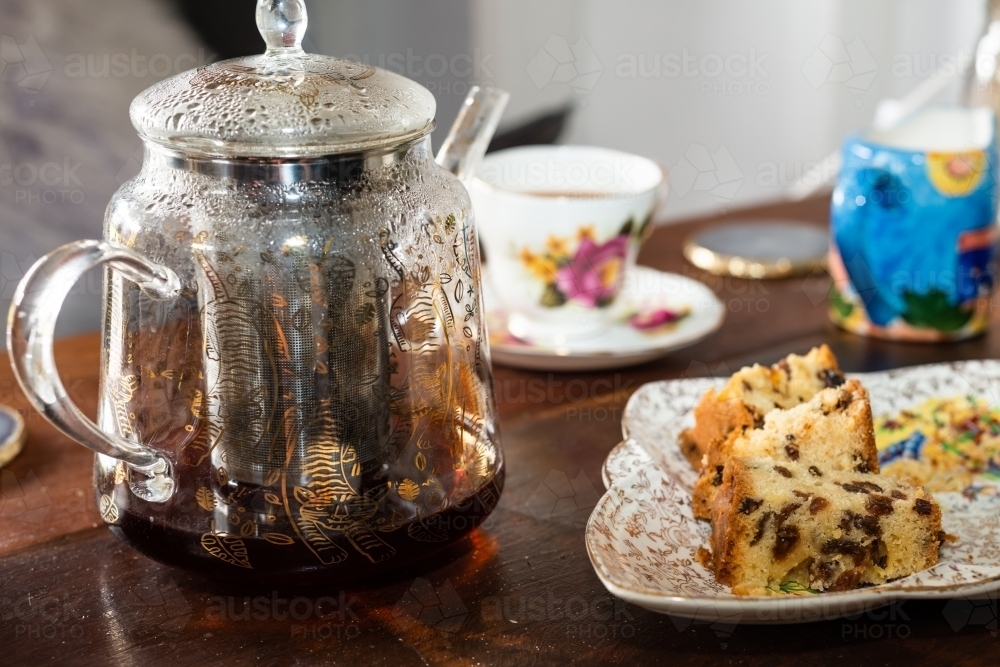 Decorative glass teapot with fine china teacup and saucer and sultana cake - Australian Stock Image