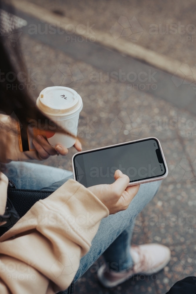 Detail of a young woman with a coffee scrolling on her phone waiting at a bus stop - Australian Stock Image
