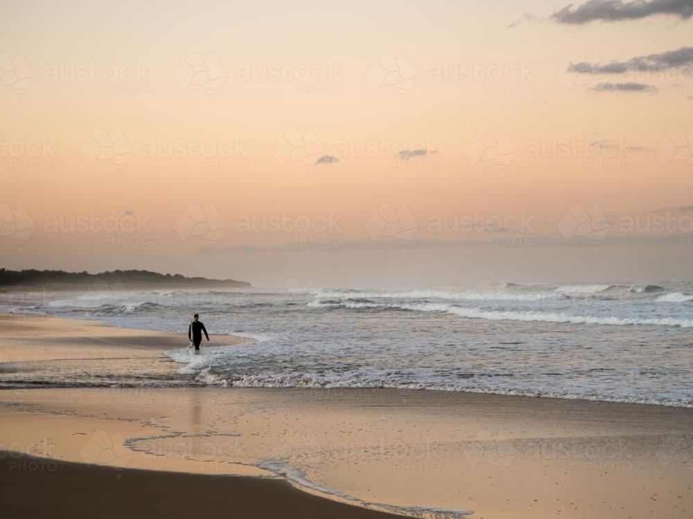 Distant lone surfer emerging from the surf at dusk - Australian Stock Image