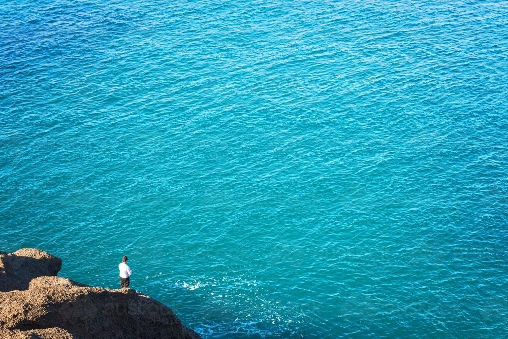 Distant person fishing off a rock beside clear blue sea waters - Australian Stock Image