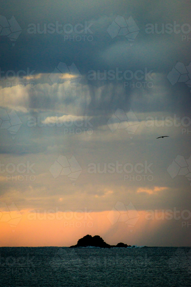 Distant rain falling over ocean at dusk with silhouette of rocky islet - Australian Stock Image