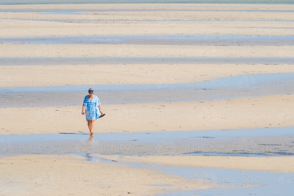 Distant view of a lady walking on a wide sandy beach with pools of water - Australian Stock Image