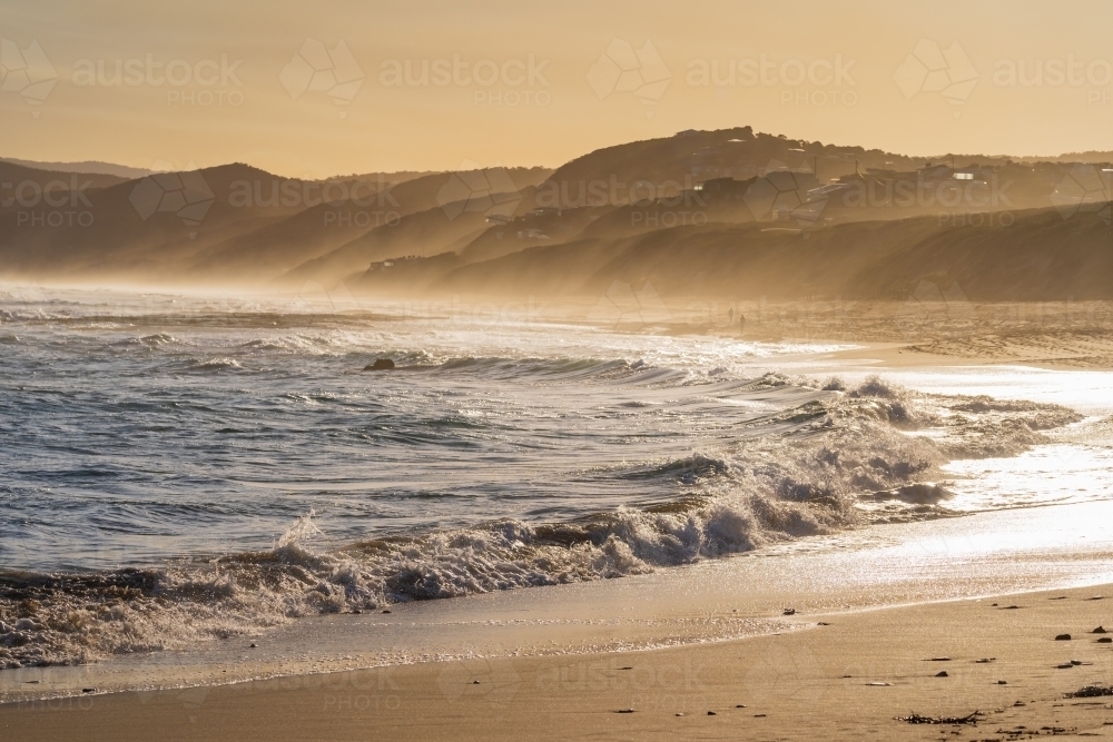 Distant view of sea mist rolling up over coastal hills above waves breaking on a beach at sunset - Australian Stock Image