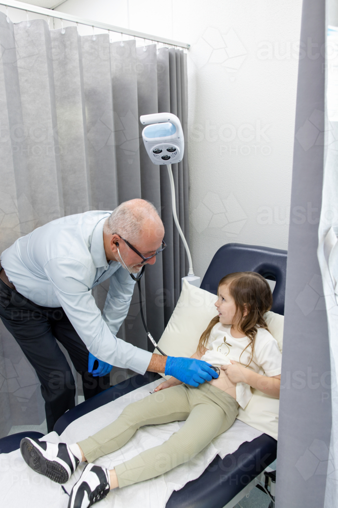 Doctor checking young girls tummy with mum standing carrying toddler son - Australian Stock Image