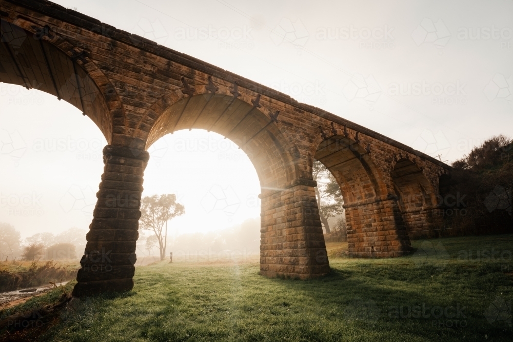 Early morning light filtering through fog under arches of historic bridge - Australian Stock Image
