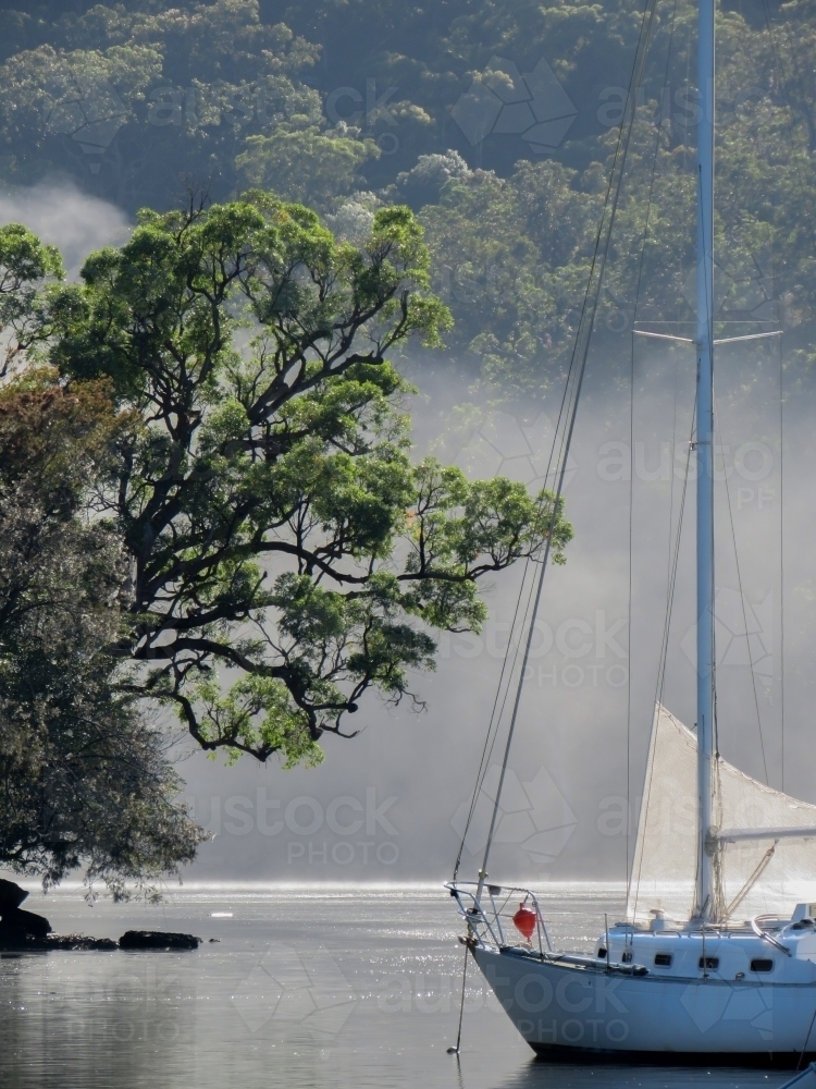 Early morning mist rising off water, with a moored yacht - Australian Stock Image
