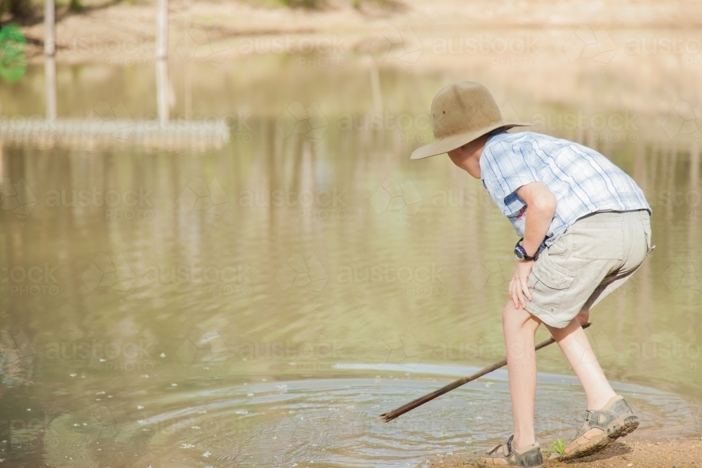 Eight year old boy splashing with stick in dam water from mud bank - Australian Stock Image
