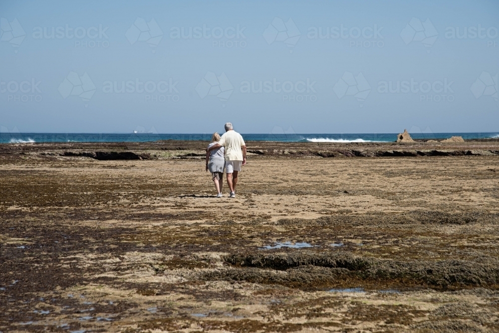 Elderly couple walking on the rocks at low tide at the beach together - Australian Stock Image