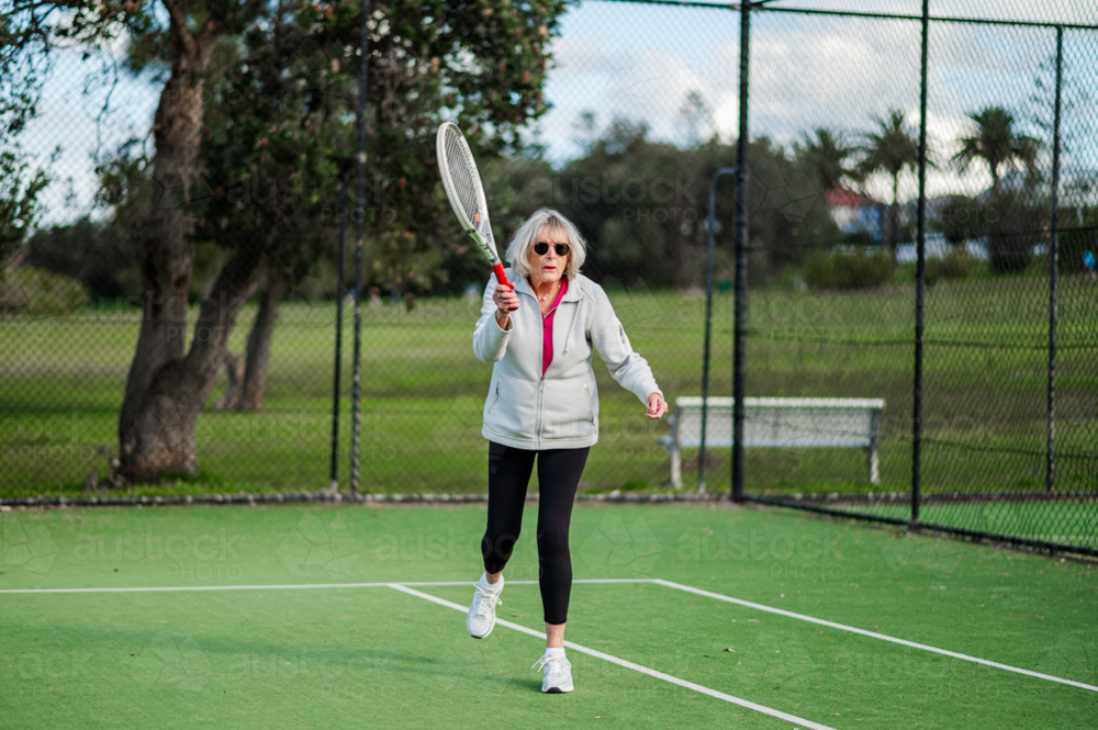 Elderly female athlete actively playing tennis on a green outdoor court - Australian Stock Image
