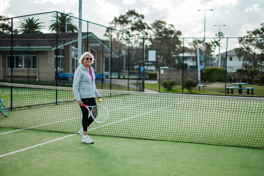 Elderly female athlete stands with a tennis racquet on a green outdoor court - Australian Stock Image