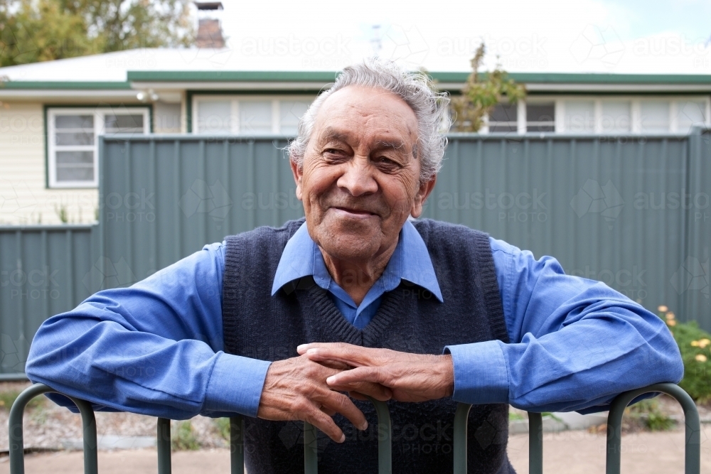 Elderly indigenous man smiling for camera while leaning on fence - Australian Stock Image