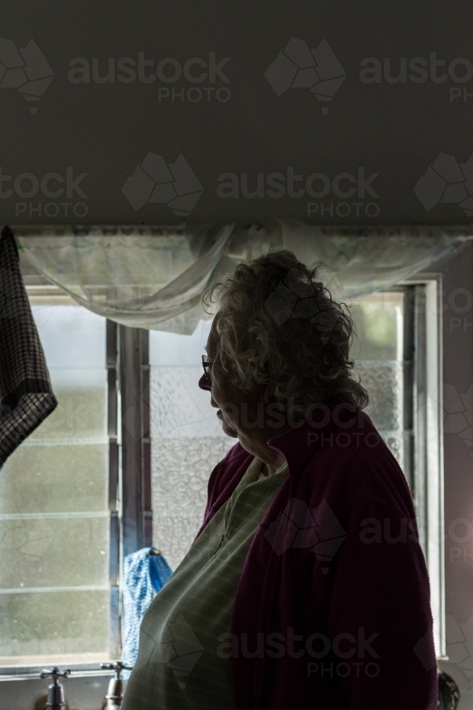 Elderly lady in gloomy room, looking out the window - Australian Stock Image