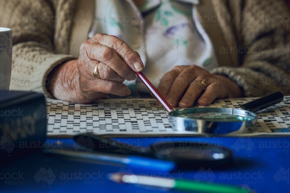 Elderly lady's hands with crossword puzzle, magnifying glass, pencil, wedding ring, and dictionary - Australian Stock Image