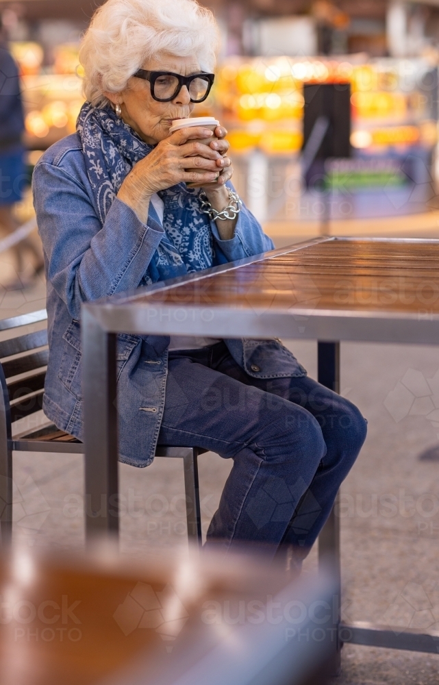 elderly lady wearing denim sitting at cafe table alone and sipping coffee in a disposable cup - Australian Stock Image