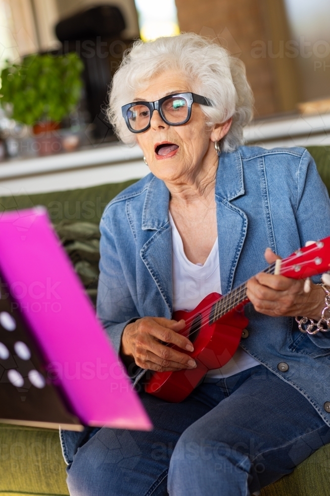 elderly lady wearing thick-rimmed glasses singing and playing ukulele - Australian Stock Image