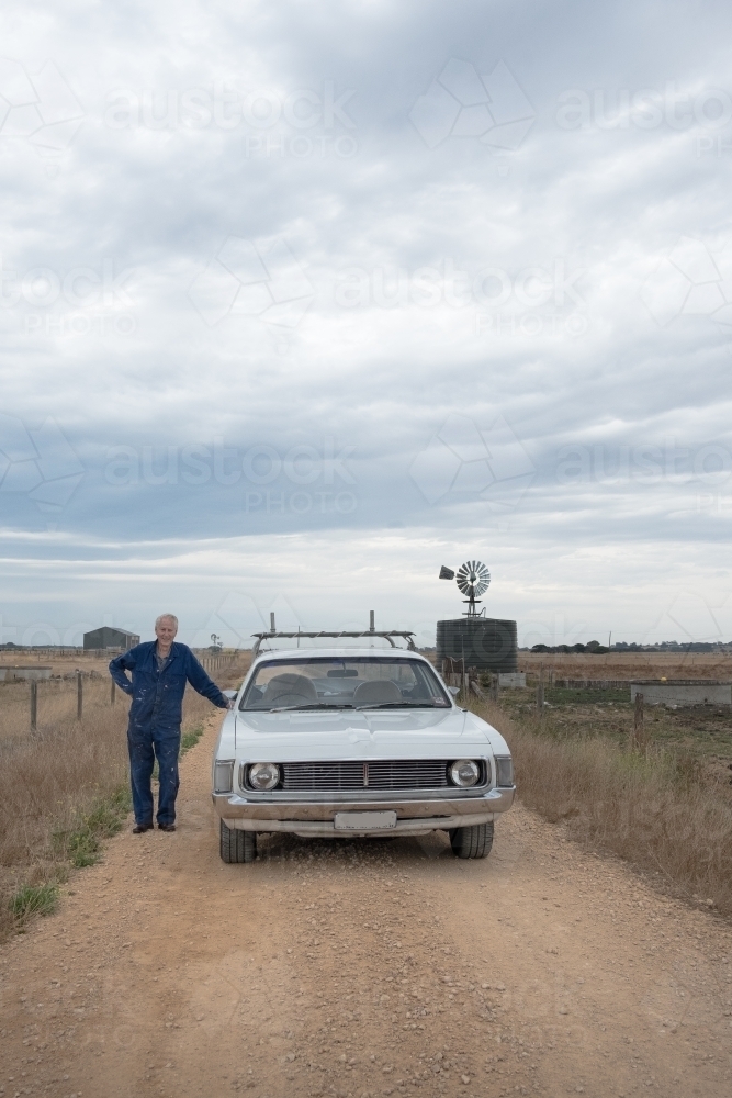 elderly man and his car on a dusty country road with a windmill. - Australian Stock Image