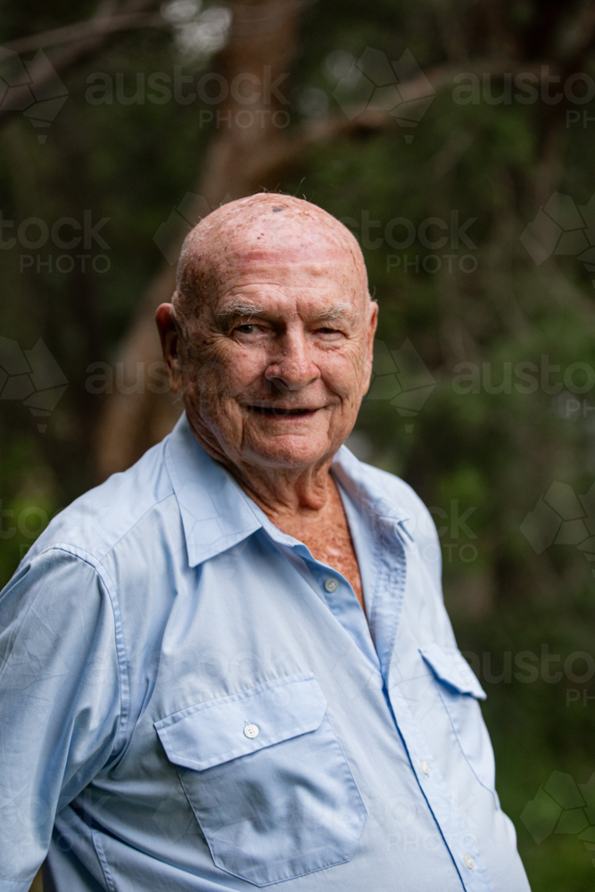 Elderly man in light blue button-up shirt standing outdoors among green trees in daylight - Australian Stock Image
