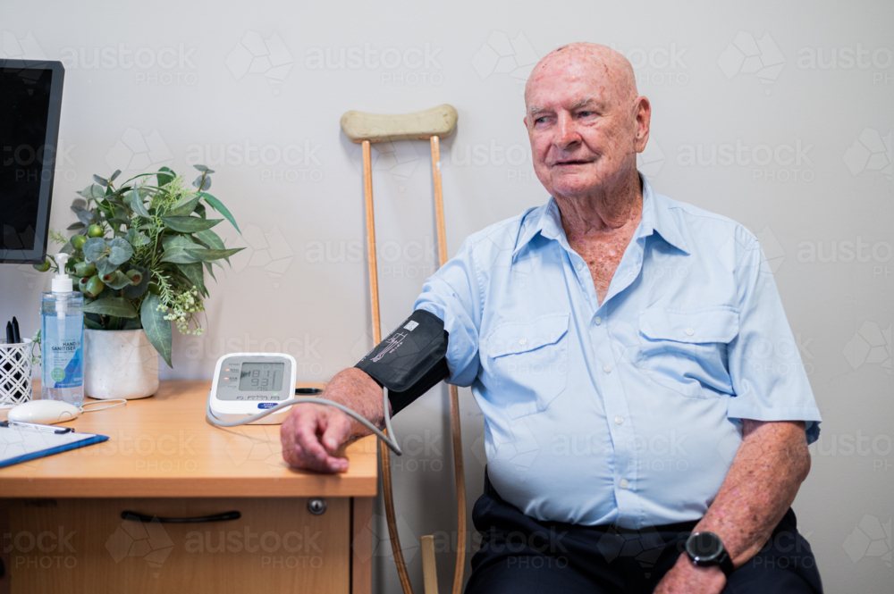 Elderly man seated near the desk with blood pressure cuff around his arm - Australian Stock Image
