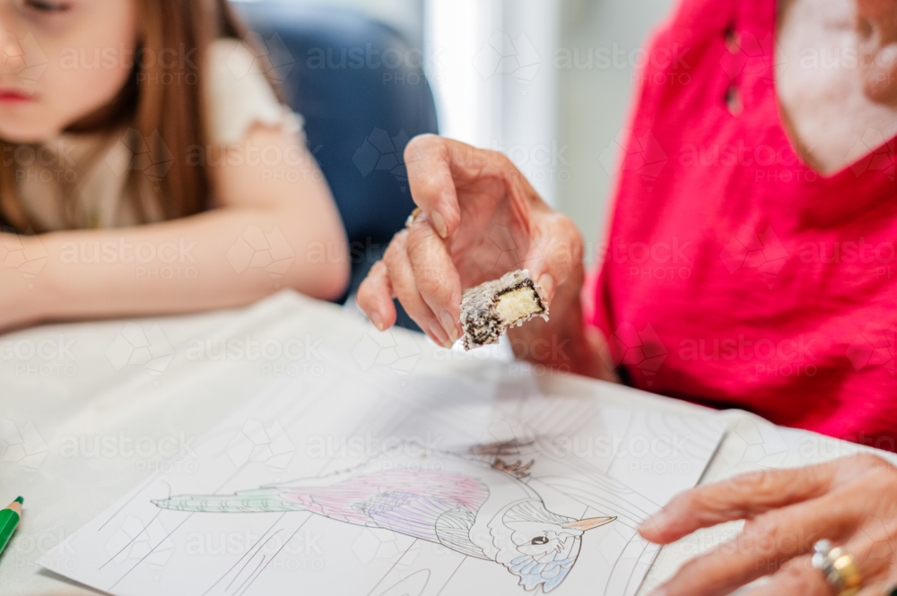 Elderly woman holding a piece of lamington cake while seated at a well-lit table - Australian Stock Image