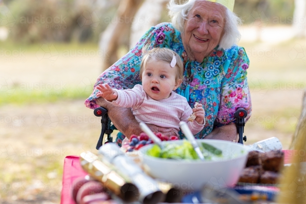 elderly woman holding baby on her lap with food on table - Australian Stock Image