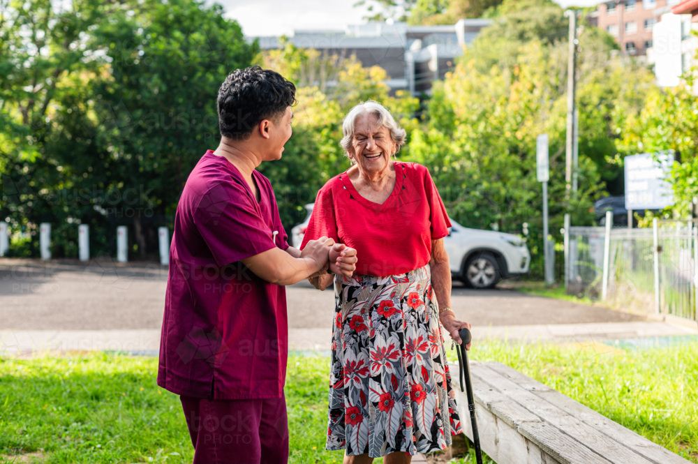 Elderly woman in a cane walking with home caregiver outdoors in grassy yard on sunny day - Australian Stock Image