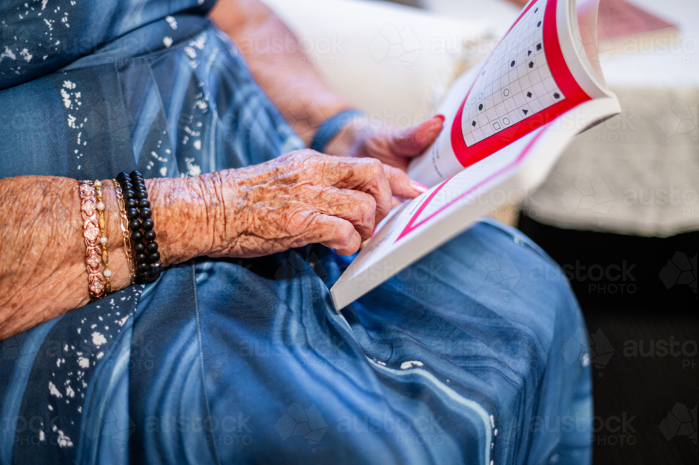 Elderly woman in blue patterned dress sitting on patterned armchair reading a book - Australian Stock Image