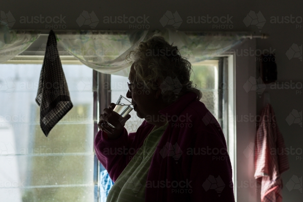Elderly woman in dim room drinking water - Australian Stock Image