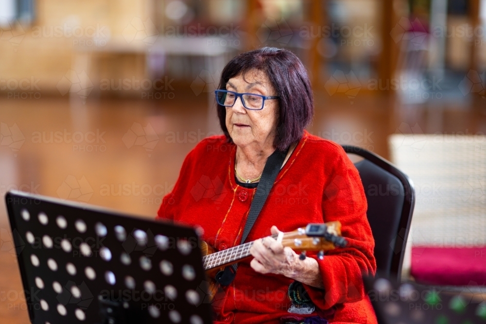 elderly woman in red playing ukulele and reading music on music stand - Australian Stock Image