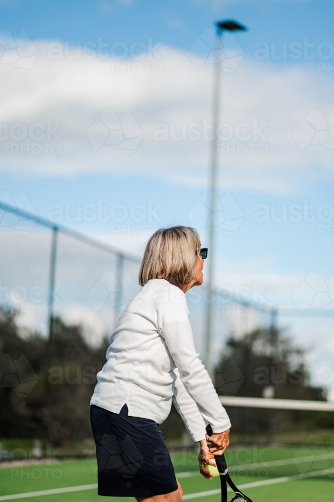 Elderly woman in sportswear prepares to hit a tennis ball on an outdoor court - Australian Stock Image