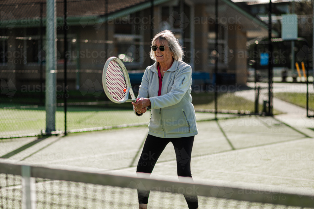 Elderly woman playing tennis, smiling and enjoying the sunny weather at an outdoor court - Australian Stock Image