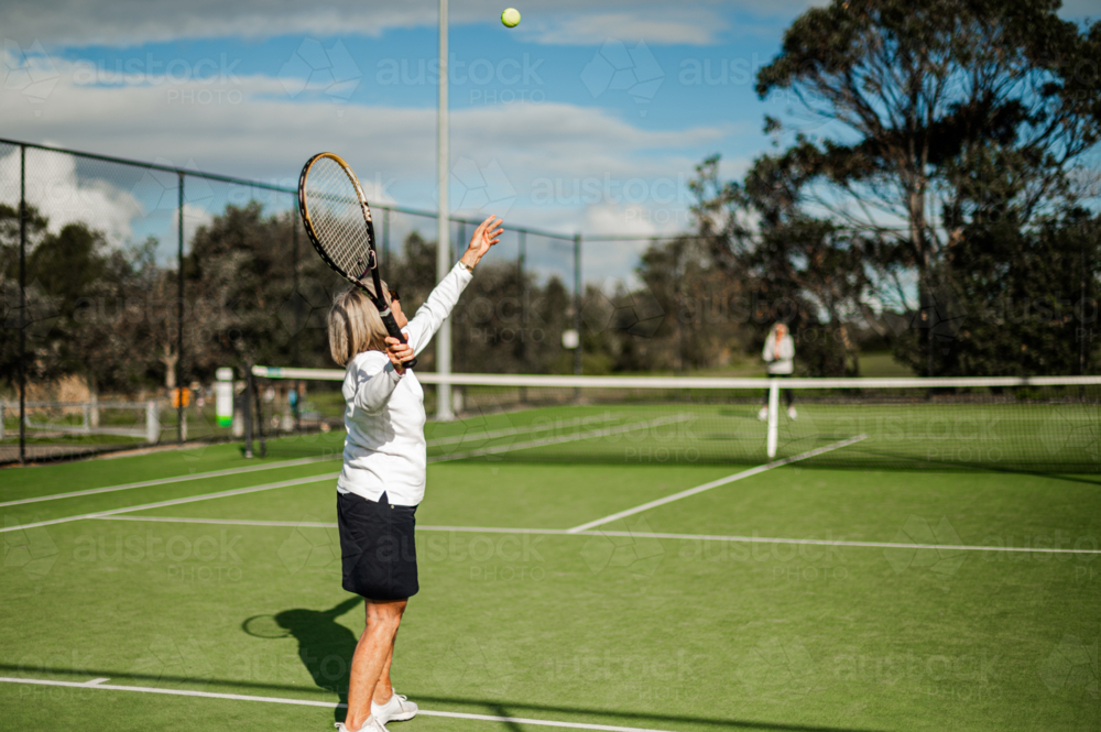 Elderly woman serves tennis ball on a bright green outdoor court, with a sunny sky - Australian Stock Image