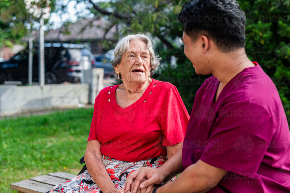 Elderly woman sitting on wooden bench with male healthcare worker sharing a conversation - Australian Stock Image