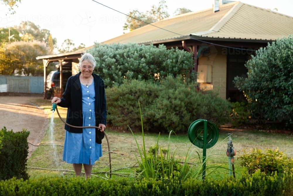 Elderly woman standing at the fence watering garden in front of house - Australian Stock Image