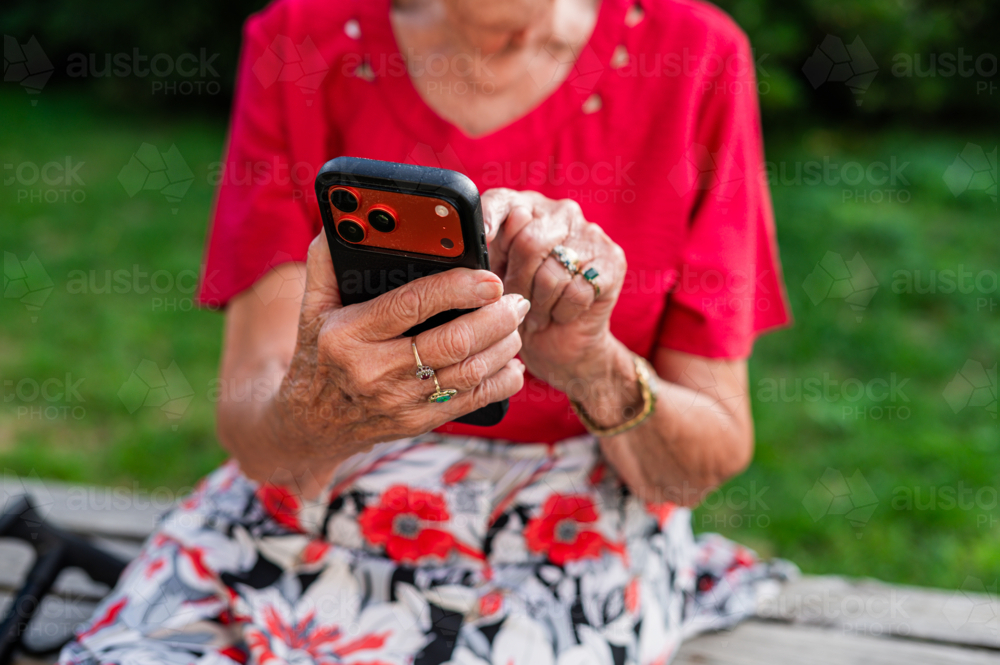 Elderly woman tapping on her phone while seated on an outdoor bench - Australian Stock Image