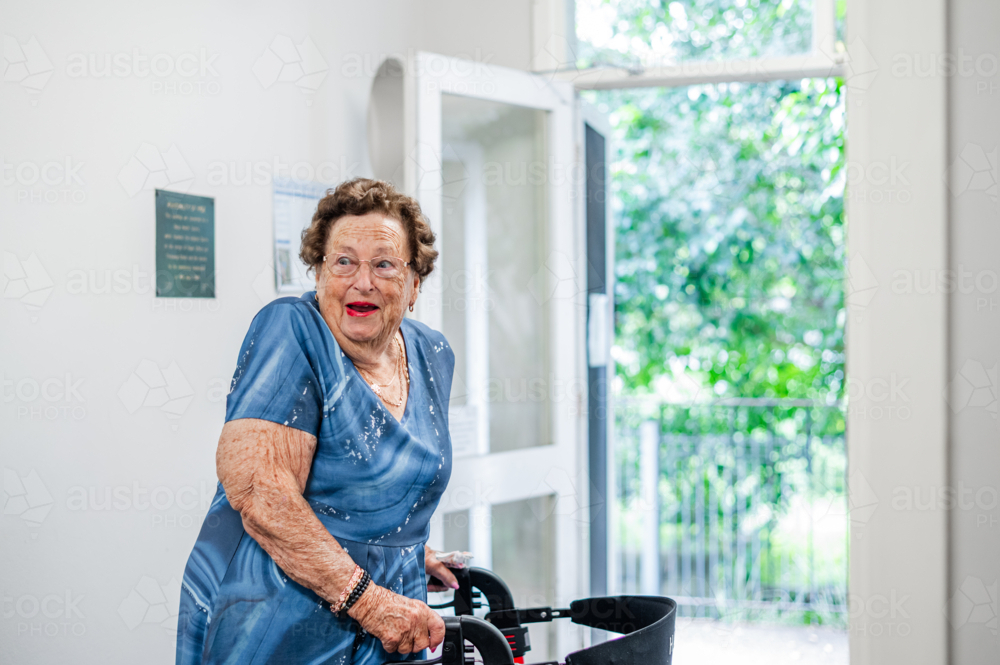 Elderly woman using her walker looking back smiling near an open door - Australian Stock Image