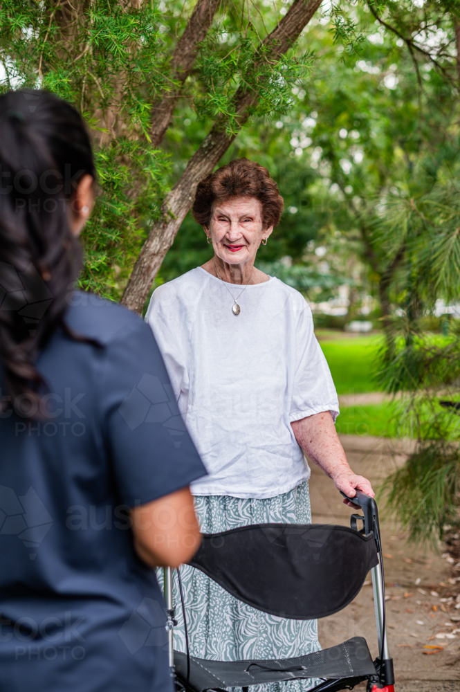 Elderly woman walking on paved footpath with her walker while care worker is approaching her - Australian Stock Image