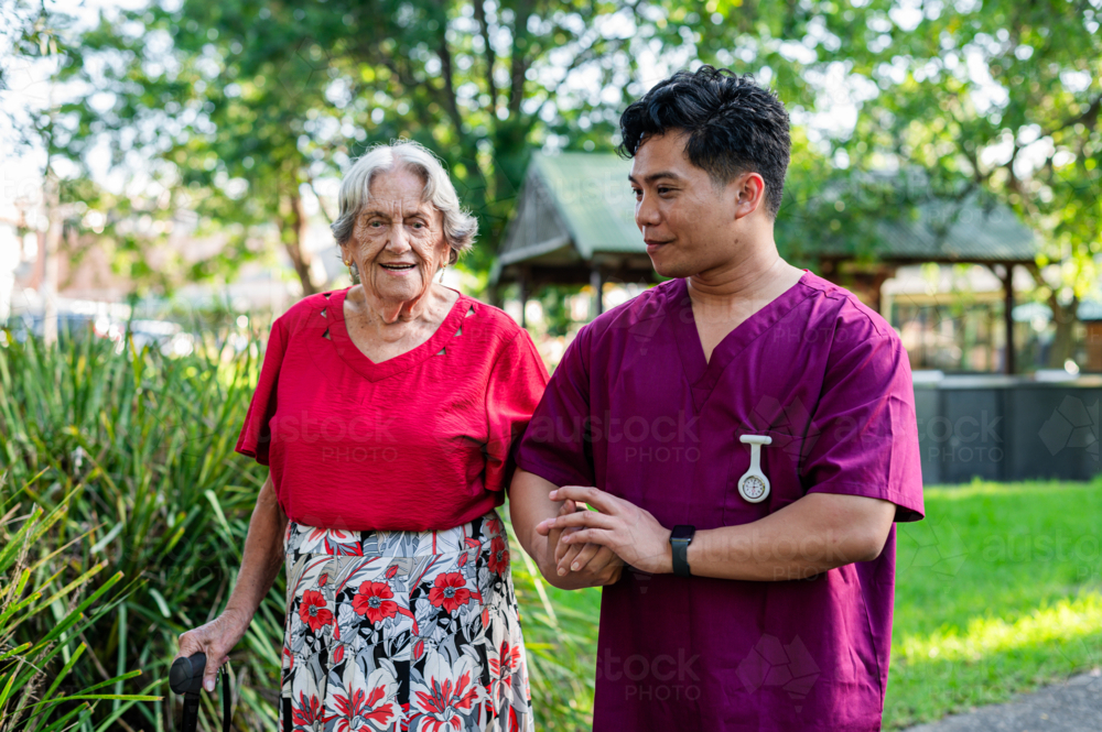 Elderly woman walking with home caregiver outdoors on sunny day - Australian Stock Image