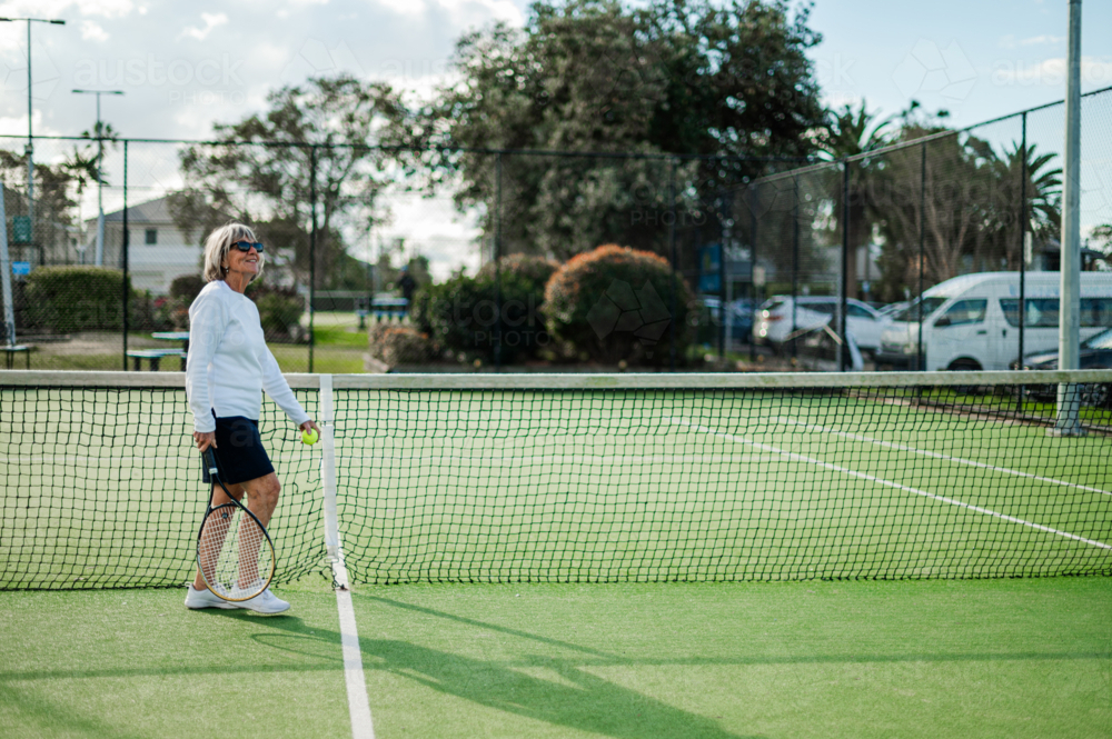 Elderly woman walks next to net, holding a racquet on an outdoor tennis court - Australian Stock Image