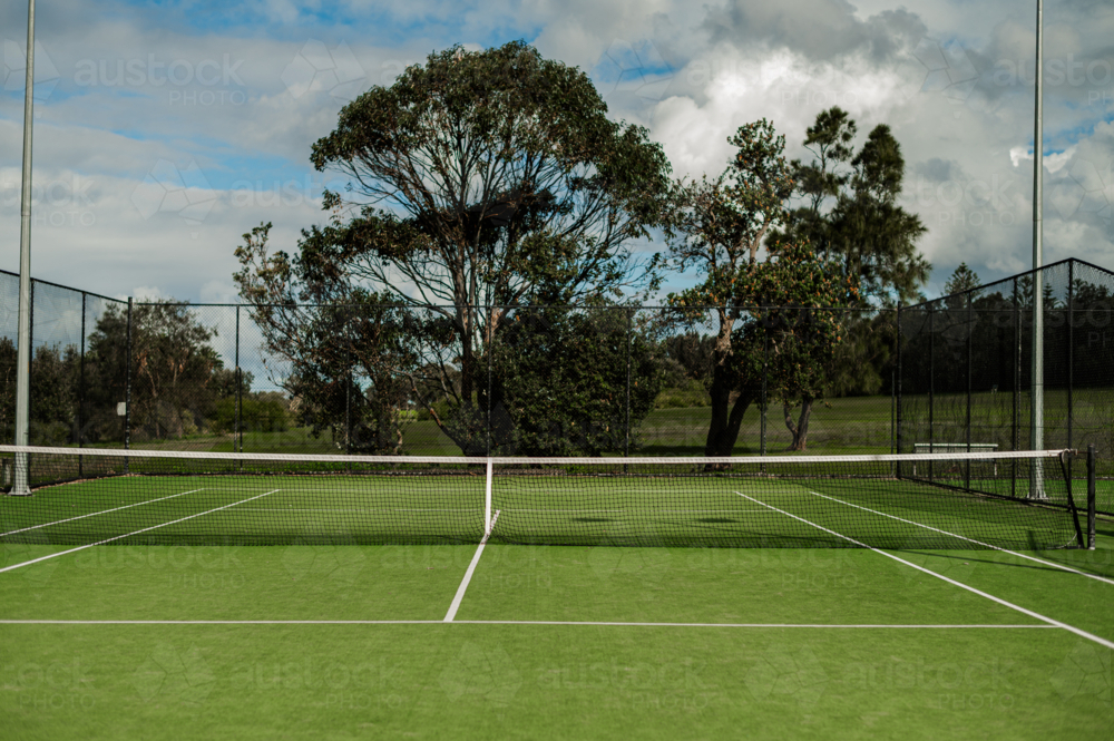 Empty tennis court features green grass and trees surrounding the area, under cloudy skies. - Australian Stock Image