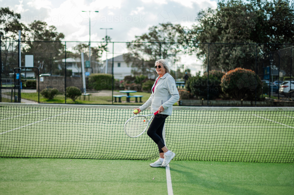 Enthusiastic woman stands by the net on an outdoor tennis court, ready to play - Australian Stock Image