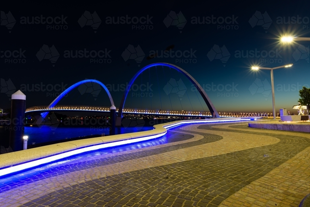 Evening view of arched bridge against blue evening sky with leading lines of blue lights and pavers - Australian Stock Image