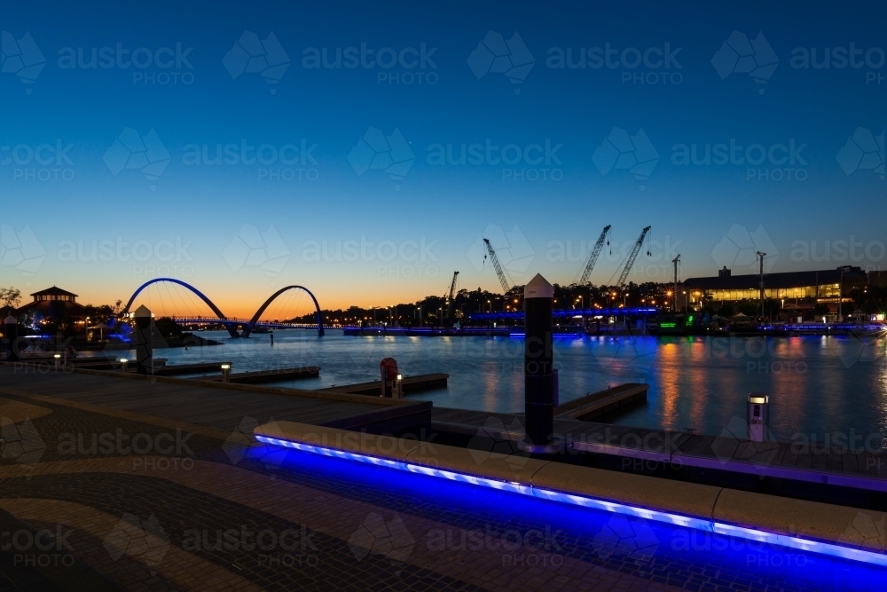 Evening view of Elizabeth Quay with arched bridge against orange glow of sunset and reflected lights - Australian Stock Image