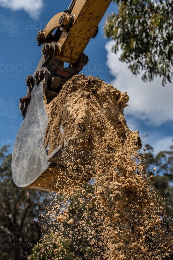 Excavator emptying bucket of dirt against blue sky - Australian Stock Image