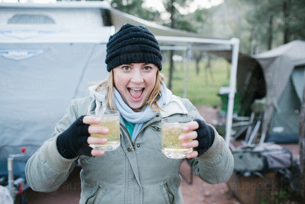 Excited young woman in beanie and gloves holding two drinks at a camp site - Australian Stock Image