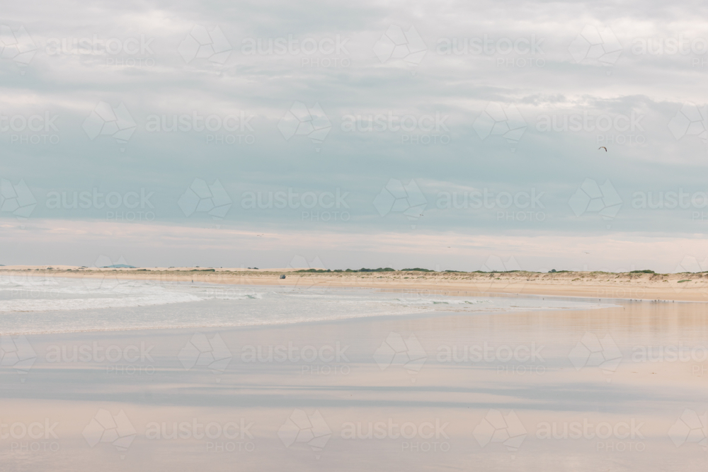 Expansive beach landscape with gentle waves under a cloudy sky and distant dunes - Australian Stock Image